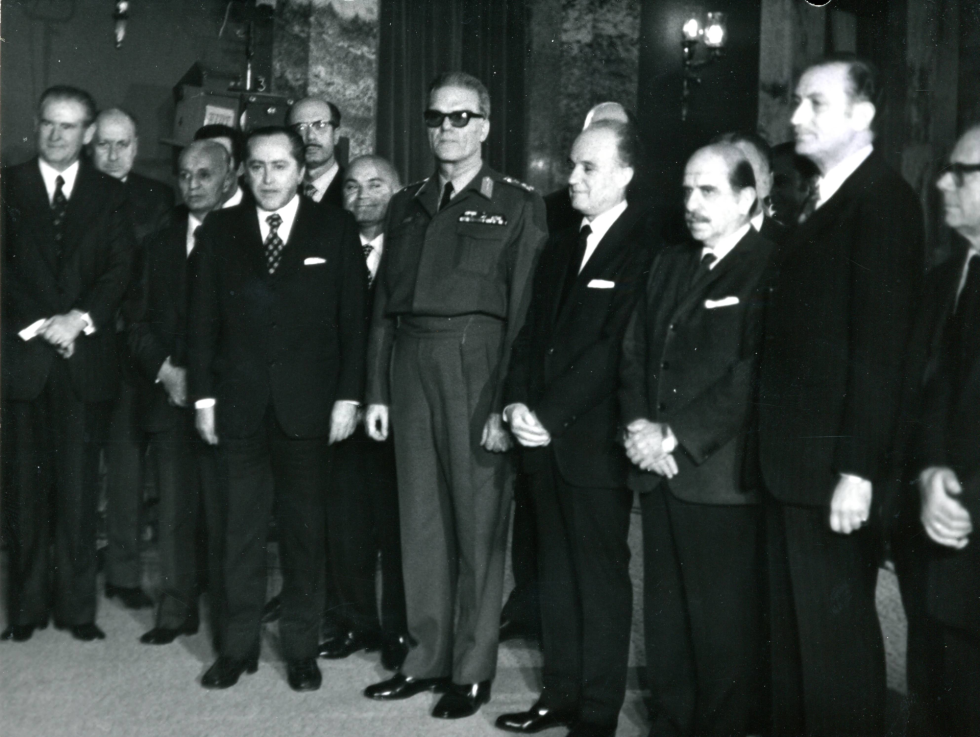 Το πρώτο κλιμάκιο του υπουργικού συμβουλίου της χούντας Ιωαννίδη (26/11/1973). Στη μέση, ένστολος ο «Πρόεδρος της Δημοκρατίας» Φίδων Γκιζίκης, στα αριστερά του ο «πρωθυπουργός» Αδαμάντιος Ανδρουτσόπουλος